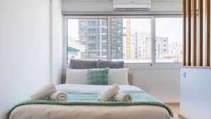 A modern bedroom with a large window, a bed with a green blanket, and a view of a construction site. - Tangerine 1-Bedroom Apartment in Larnaca (Larnaca)