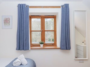 Una stanza con una finestra a cornice di legno, tende azzurre, una piccola pianta in vaso sul davanzale, un asciugamano bianco arrotolato su un letto blu e un quadro appeso al muro.