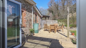 Une terrasse avec un sol en pierre, une table et des chaises en bois, et une poubelle verte.