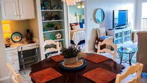 A dining area with a table set for four, surrounded by white chairs, a chandelier, and a television on a stand. - Seville 602: Cozy 2-Bedroom Ocean View Condo (South Padre Island)