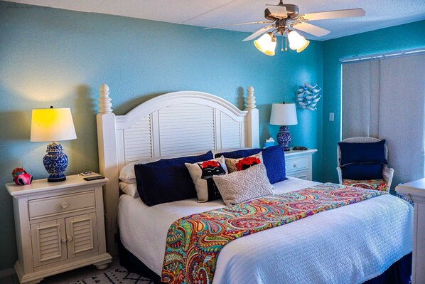 A bedroom with a white bed, blue walls, a ceiling fan, and a nightstand with a lamp.
