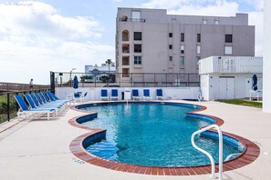 A swimming pool with blue lounge chairs and a brick border, surrounded by a fence and a building in the background.