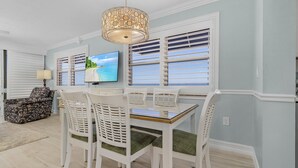 A dining area with a table and chairs, a television, and a view through the window blinds. - Beach Villas 706 Top-floor Condo W/ Gulf Views, Pool 2 Bedroom Condo (Fort Myers Beach)