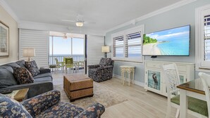 A spacious living room with a large TV, comfortable seating, and a view of the ocean. - Beach Villas 706 Top-floor Condo W/ Gulf Views, Pool 2 Bedroom Condo (Fort Myers Beach)