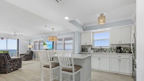 A modern kitchen with white cabinetry, a dining area with floral chairs, and a large window overlooking the ocean. - Beach Villas 706 Top-floor Condo W/ Gulf Views, Pool 2 Bedroom Condo (Fort Myers Beach)