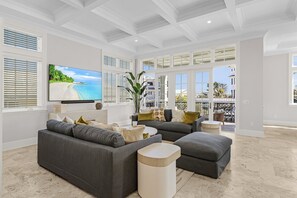 A spacious living room with a large flat-screen TV, grey sofas, and a view of the ocean through large windows. - 260 Coastal Luxe Retreat: 5bd/4.5ba, Pool & Spa, Water Views 5 Bedroom Home (Fort Myers Beach)