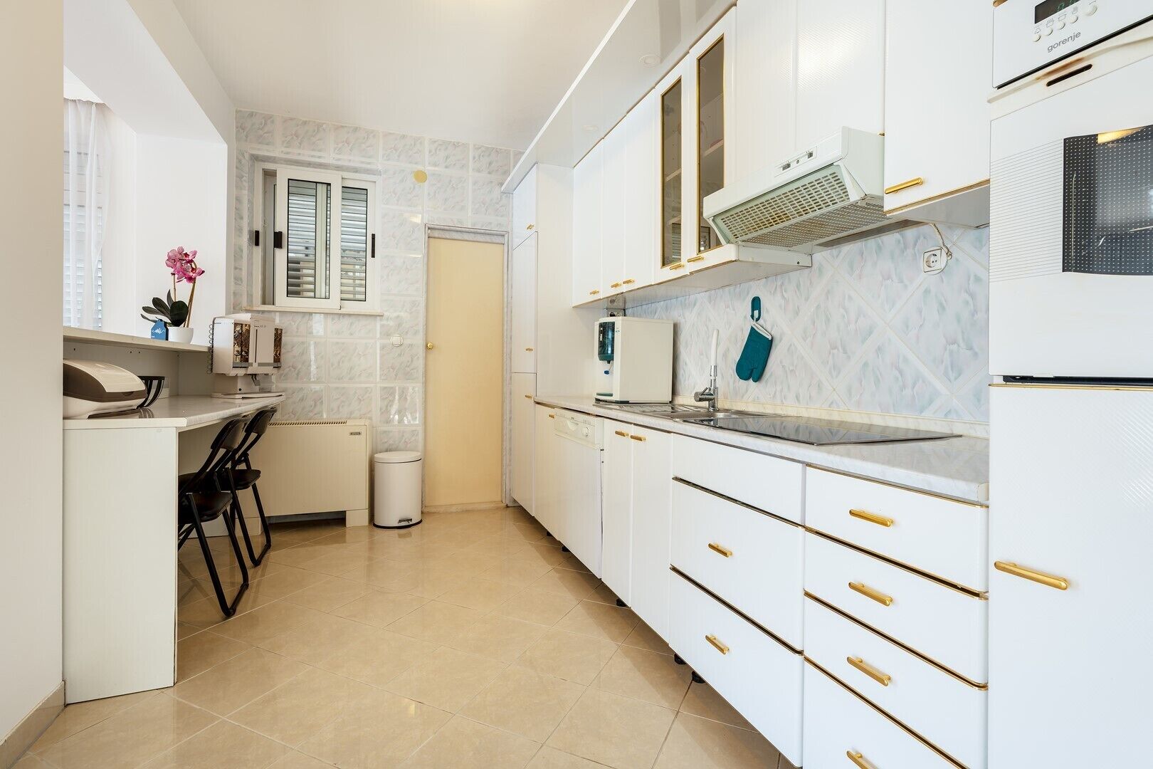 Een moderne keuken met witte kasten, een inbouwoven en een raam met jaloezieën.