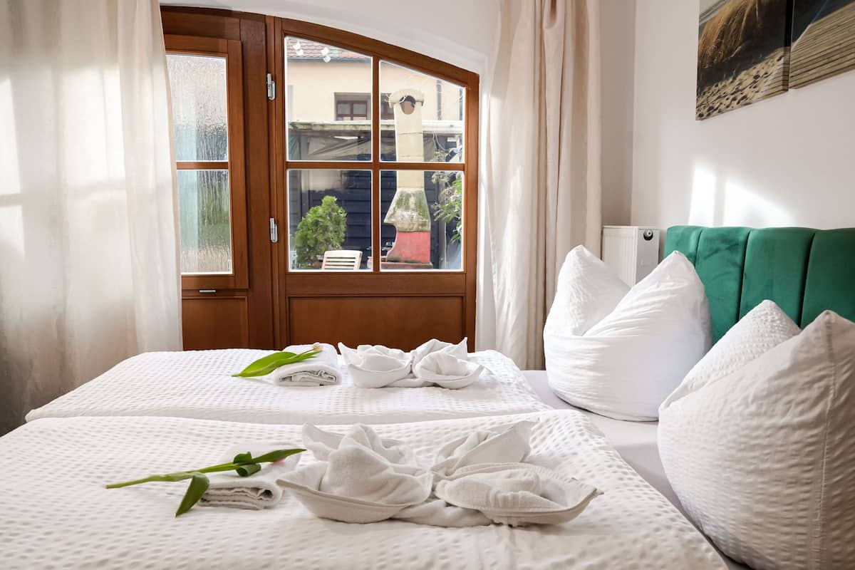 A hotel room with two beds, a green upholstered headboard, white pillows, and a window with a view of buildings and greenery.