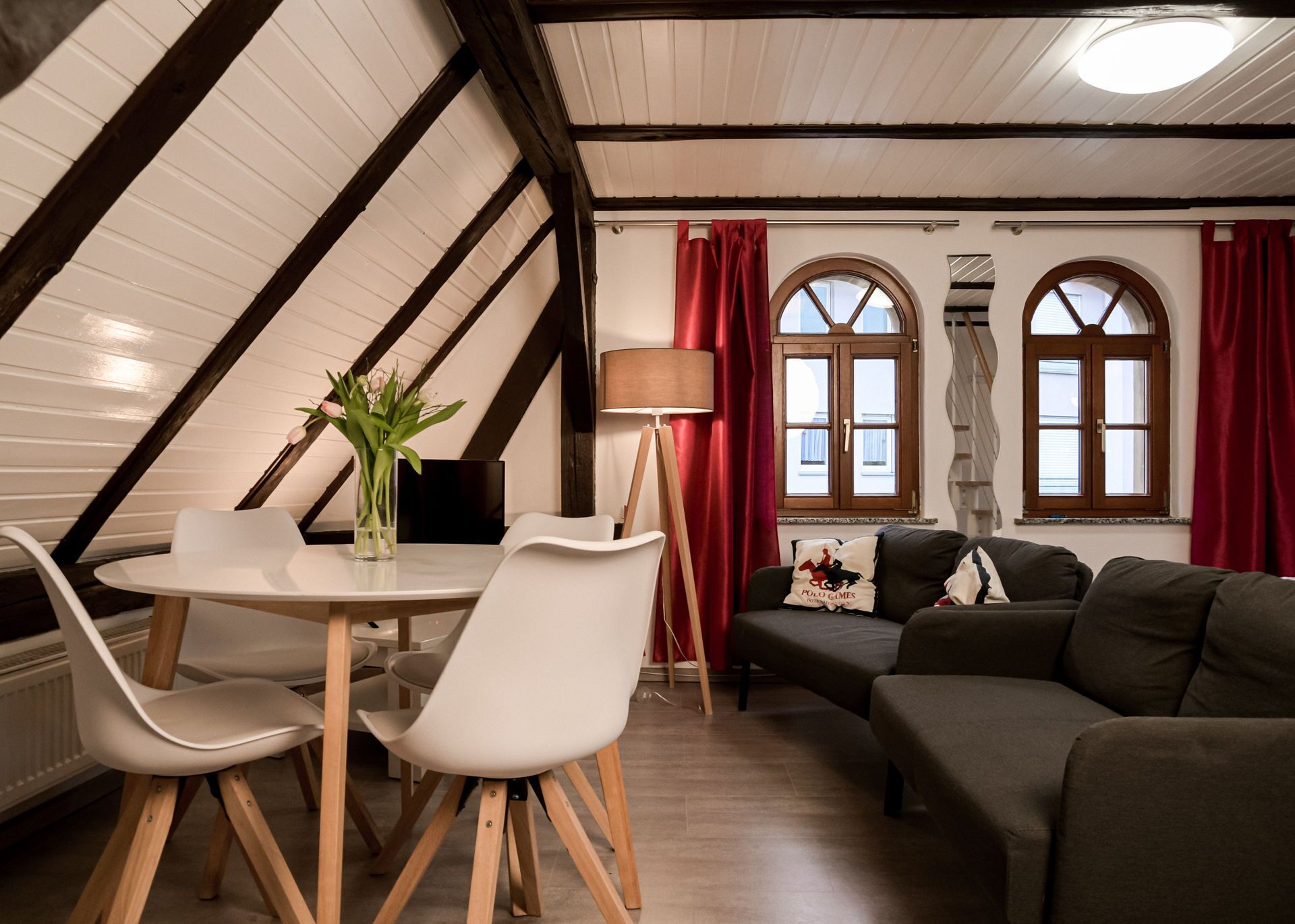 A cozy loft-style room with a dining area, a sofa, and two windows with red curtains.