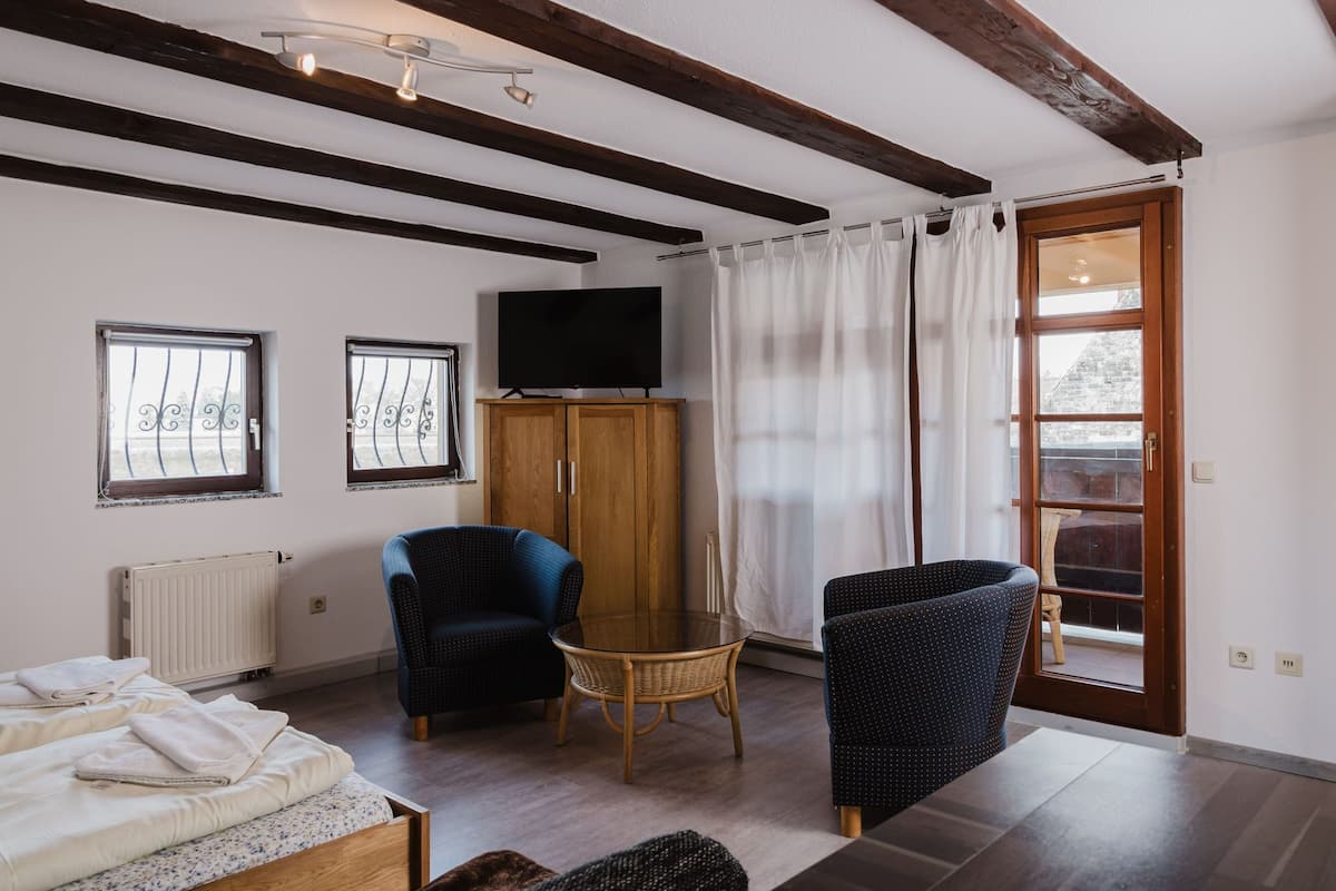 A room with a wooden ceiling, a television on a cabinet, two chairs, a small table, and a bed with towels.