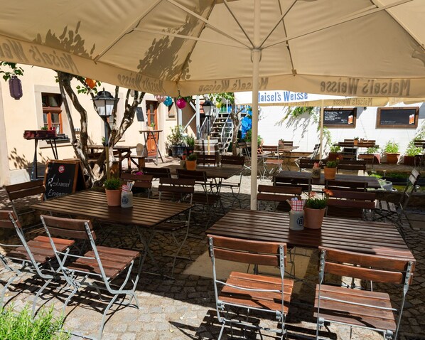 An outdoor café with wooden tables and chairs, potted plants, and a large umbrella providing shade. - Pension Trobischhof (Dresden)