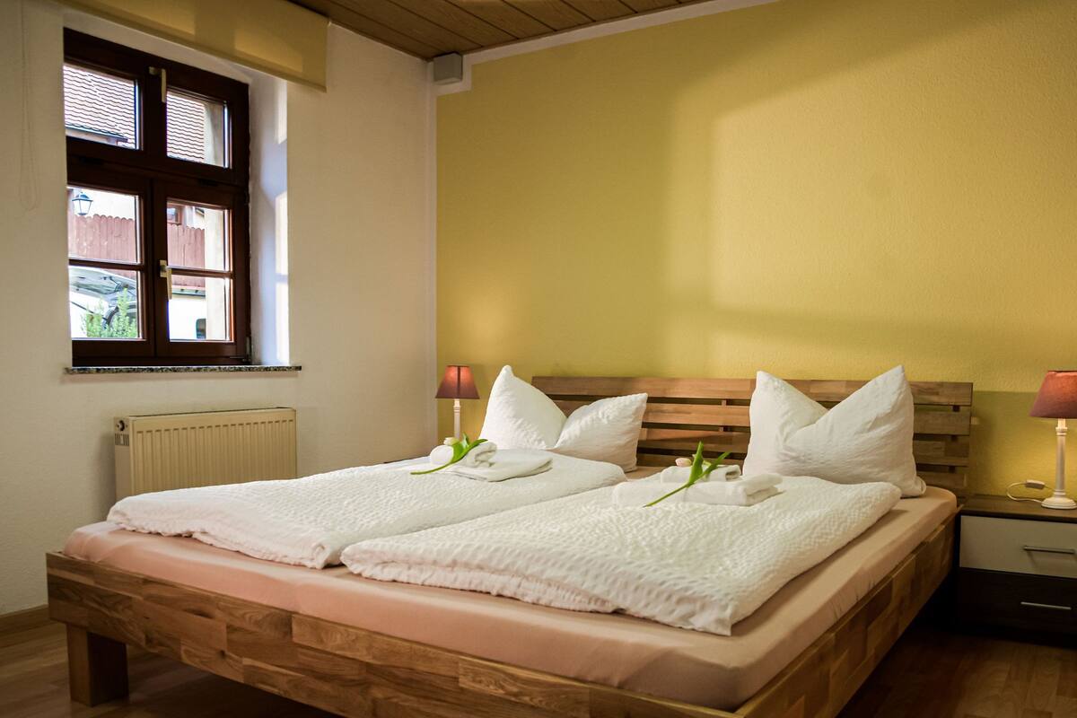A double bed with white linens and pillows, a wooden headboard, a bedside table with a lamp, and a window with blinds.