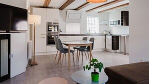 Una cocina moderna con armarios blancos, un comedor con una mesa redonda y sillas, y una sala de estar con un sofá marrón y una planta verde.