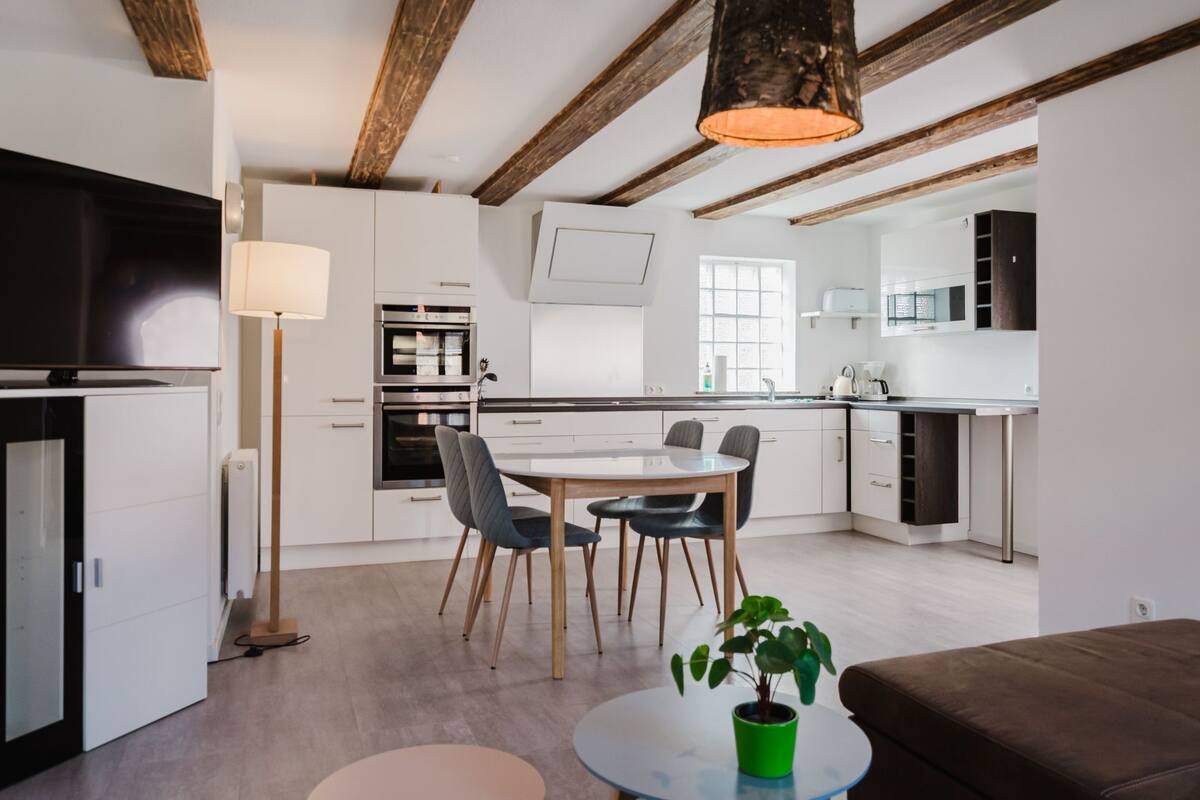 A modern kitchen with white cabinets, a dining area with a round table and chairs, and a living area with a brown sofa and a green plant.