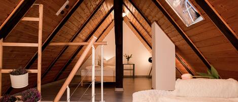A cozy attic bedroom with a sloped ceiling, wooden beams, a skylight, a ladder, and a bed with a white blanket.