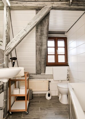 A bathroom with a rustic aesthetic, featuring exposed wooden beams, a white tiled wall, a wooden shelf with toiletries, a toilet, and a window. - Pension Trobischhof (Dresden)