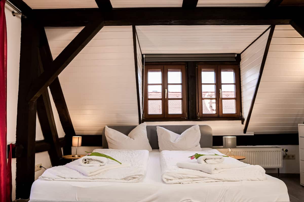 A bedroom with a large bed, two windows, wooden beams, a radiator, and a nightstand with a lamp.