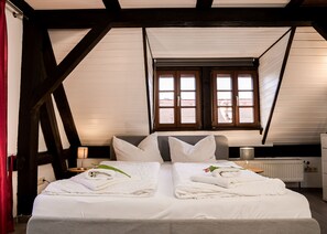A bedroom with a large bed, two windows, wooden beams, a radiator, and a nightstand with a lamp. - Pension Trobischhof (Dresden)