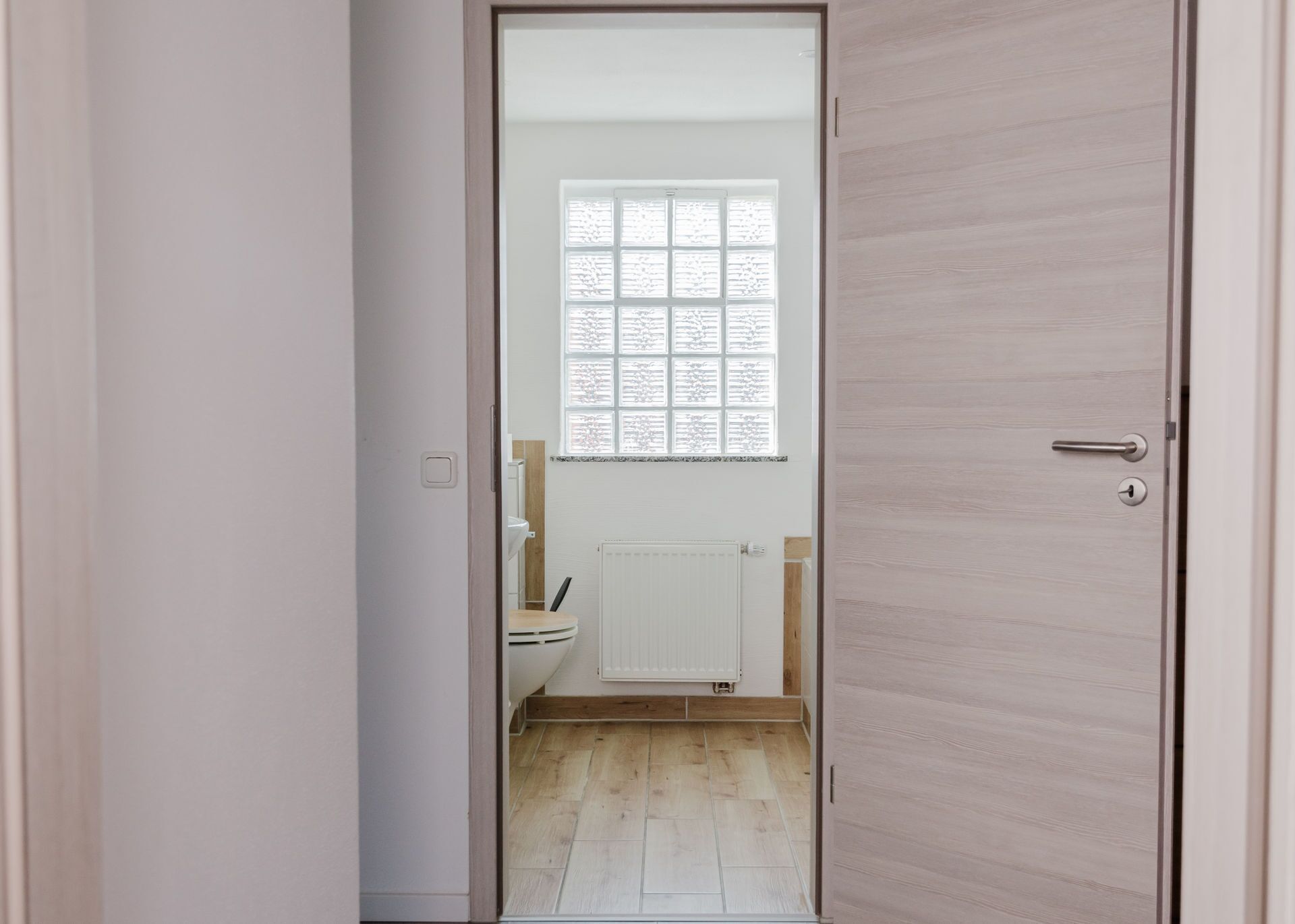 En smal gang, der fører til et badeværelse med et vindue, en radiator og et toilet.
