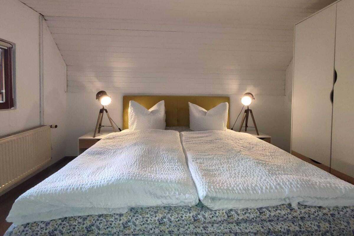 A twin bed with a white quilt, two bedside lamps, and a wooden headboard in a room with white walls and a sloped ceiling.