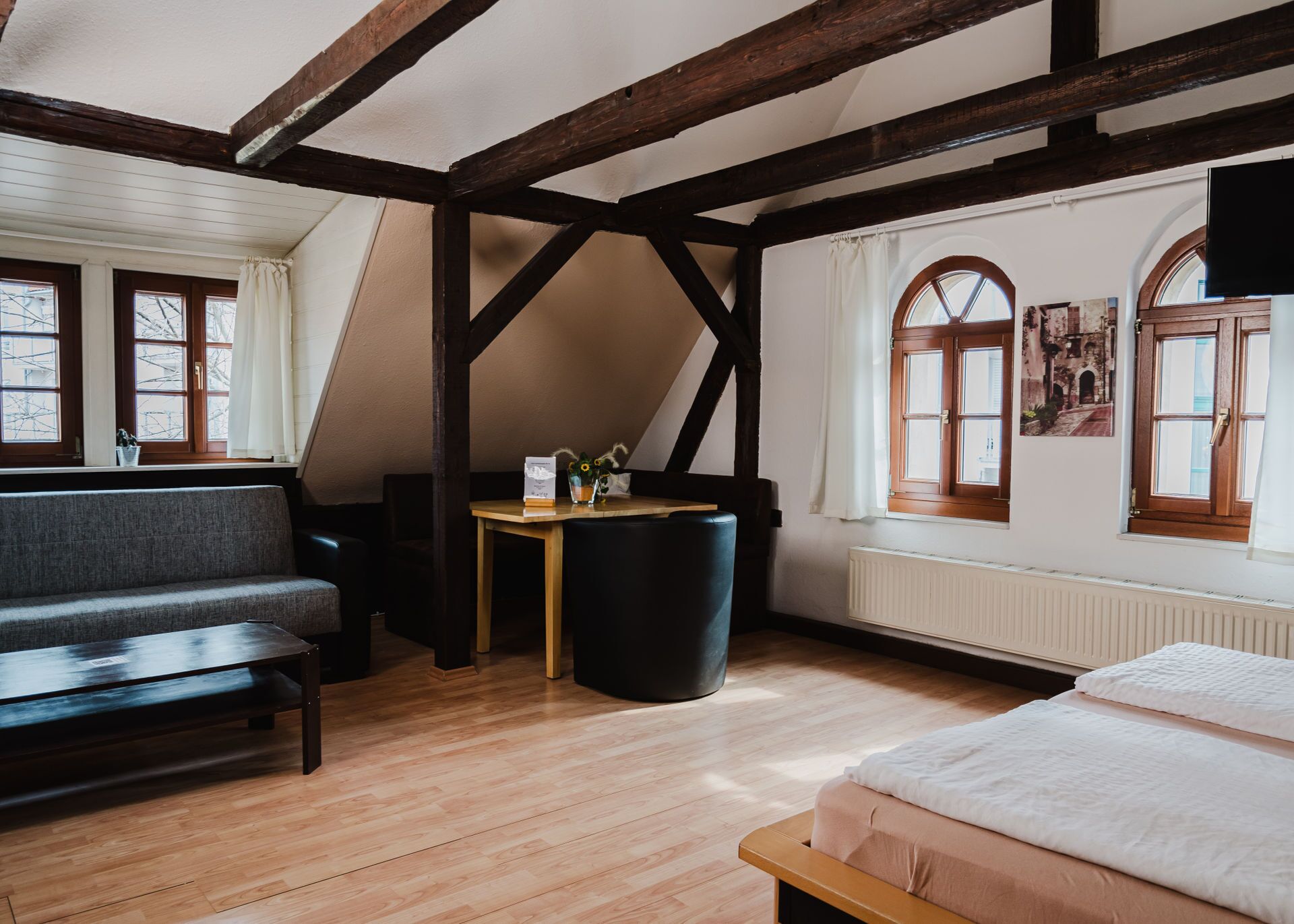 Ein Zimmer mit einem Schlafplatz in der Mansarde, einer Couch, einem kleinen Tisch mit einer Vase und einem Fenster mit Vorhängen.