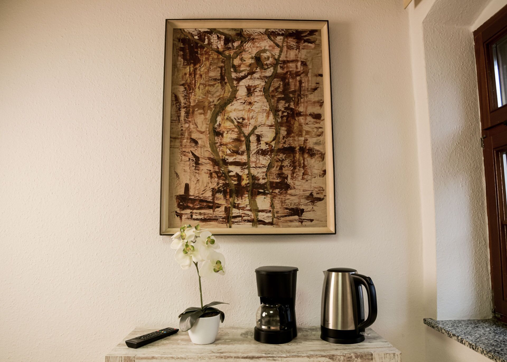 A framed abstract painting on a wall, a white orchid in a pot, a black coffee maker, and a silver kettle on a marble surface.