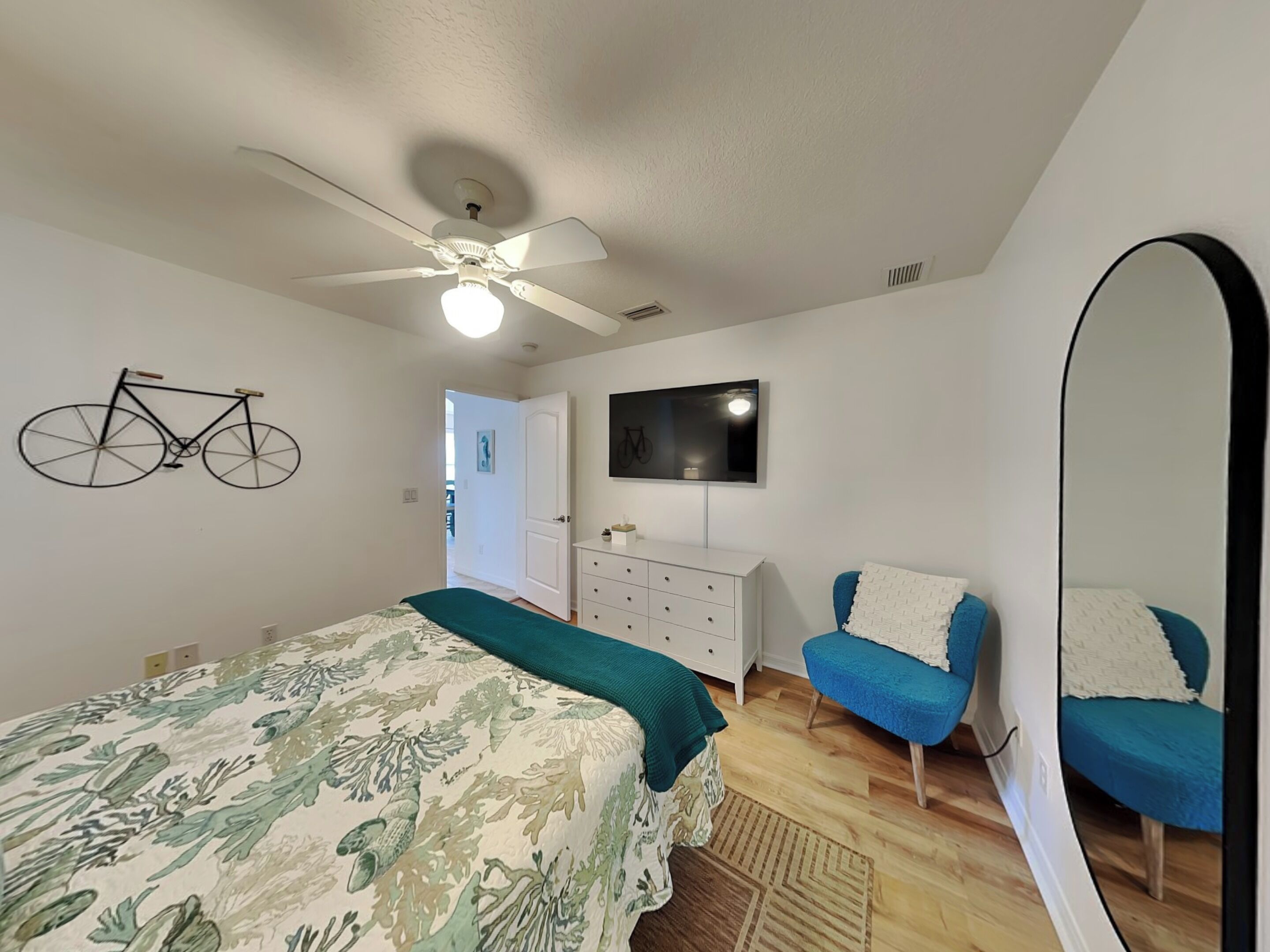 A bedroom with a bed, a chair, a dresser, a mirror, a ceiling fan, and a wall-mounted TV.