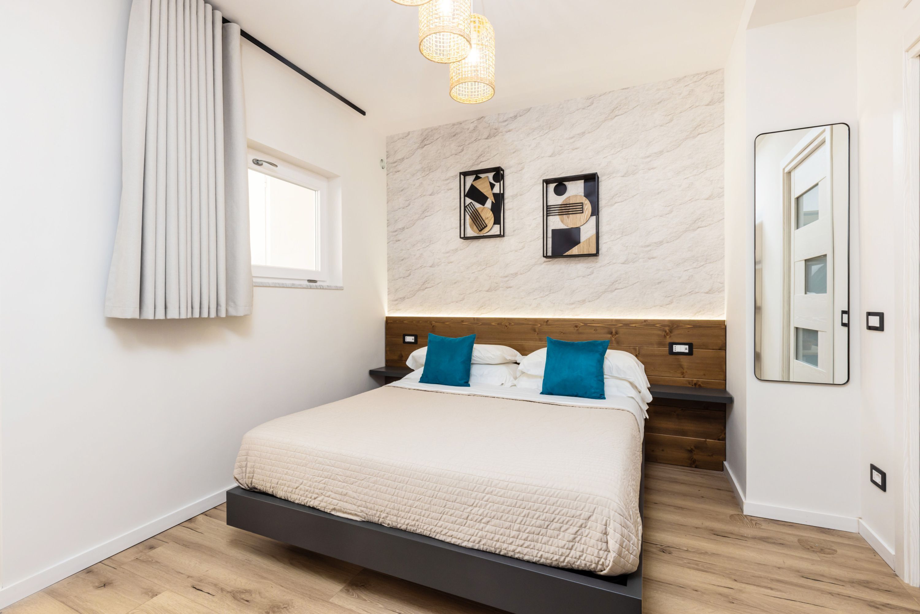 Une chambre moderne avec un lit, une tête de lit en bois, des tables de chevet, une fenêtre avec des rideaux et des œuvres d’art murales.