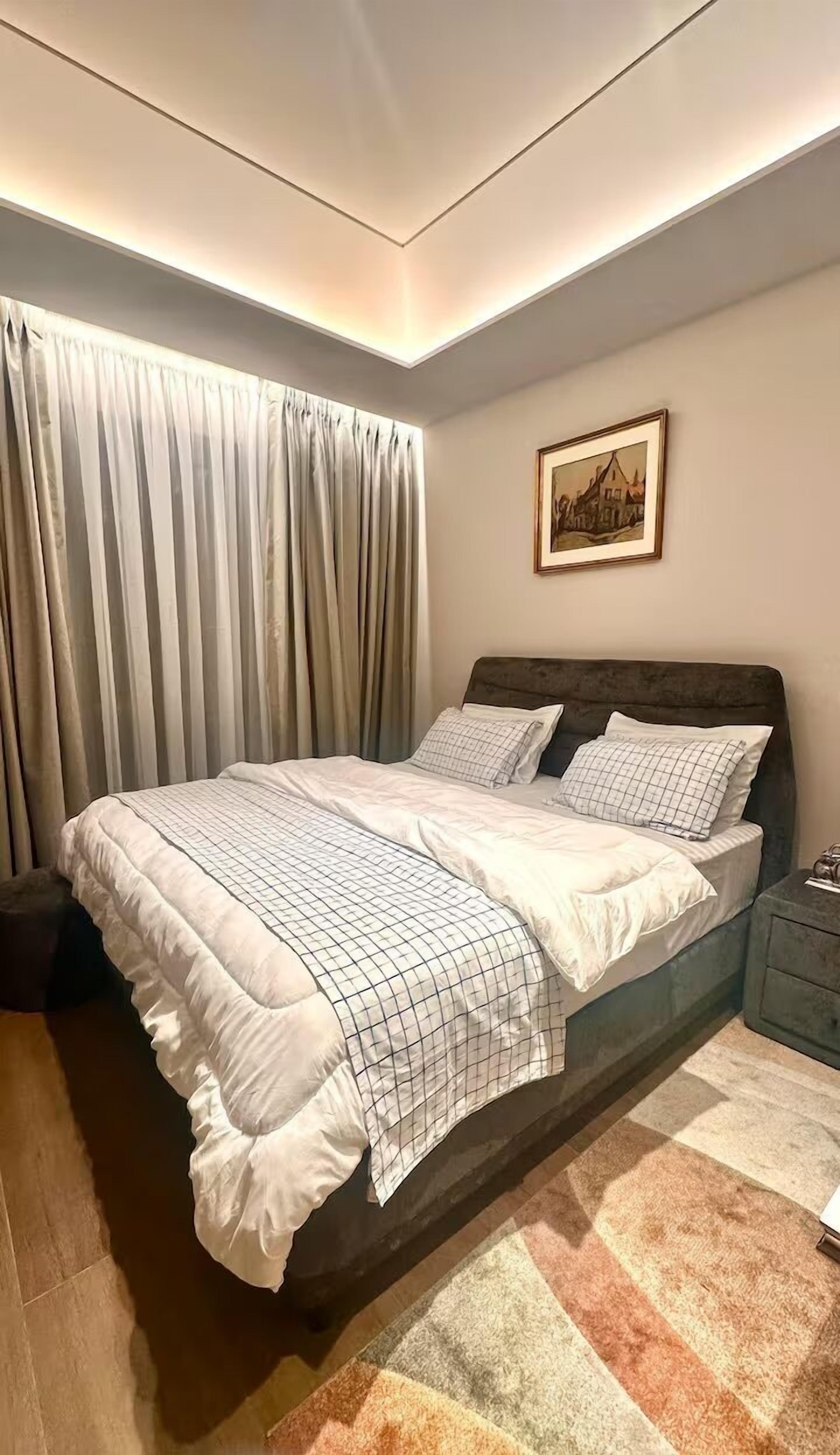 A bedroom with a bed, curtains, a nightstand, a framed picture, and a carpet.