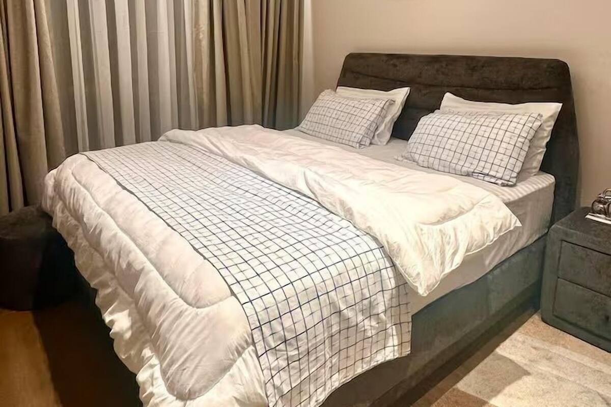 A bedroom with a bed, curtains, a nightstand, a framed picture, and a carpet.