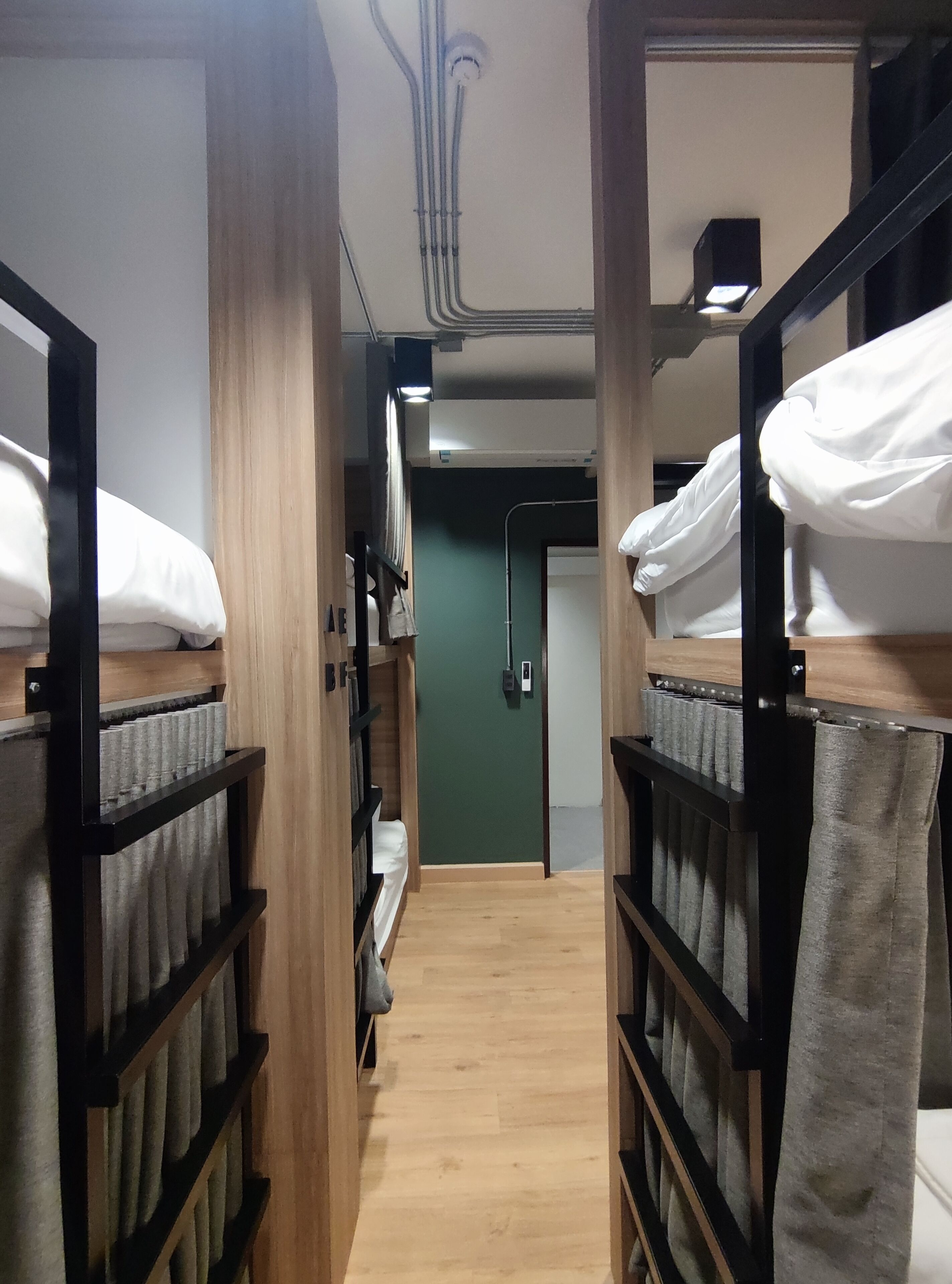 A narrow corridor with bunk beds on either side, wooden flooring, and a green wall with a door at the end.