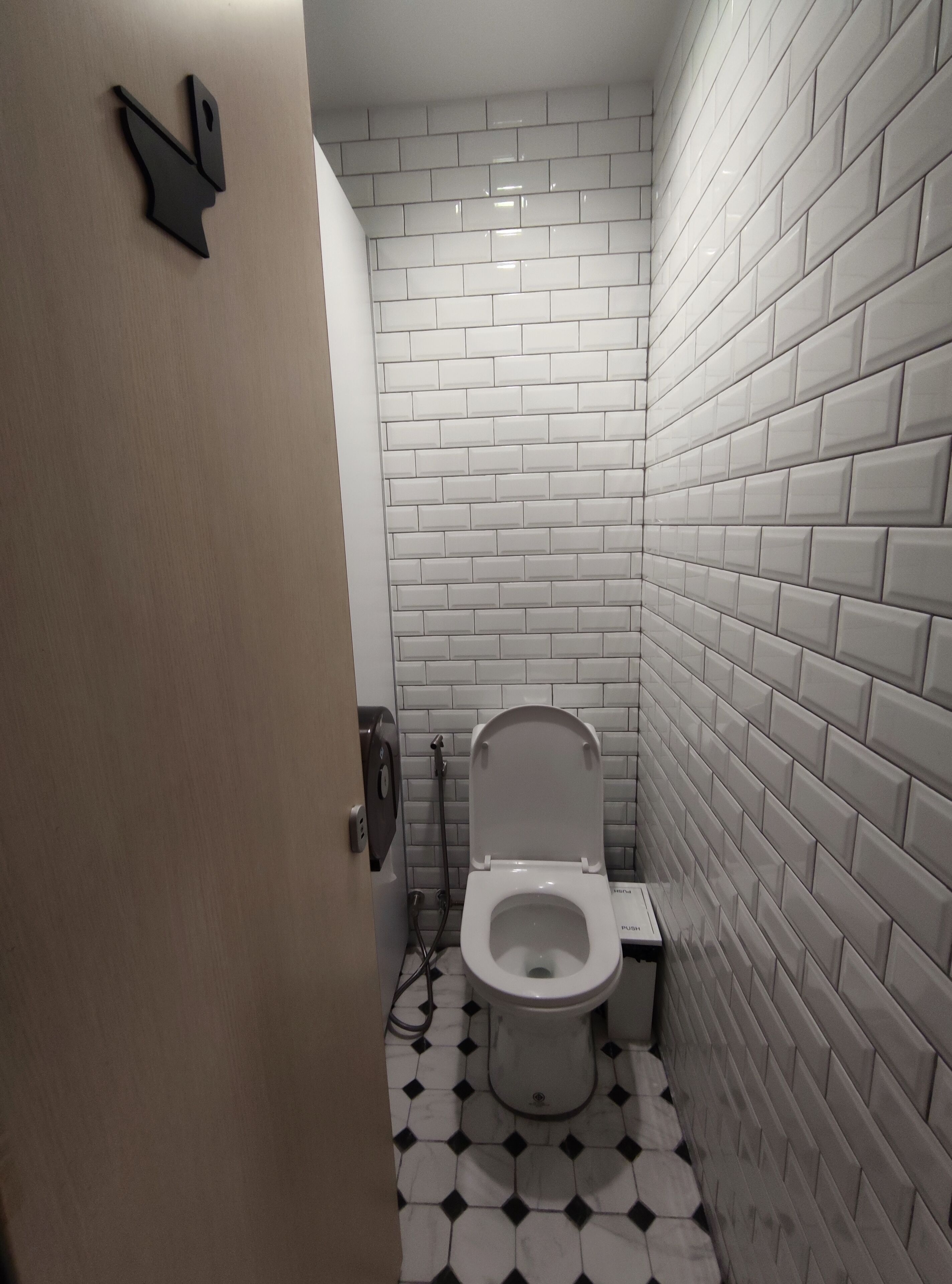 A small, narrow bathroom with white subway tile walls and a black and white patterned floor. A toilet is visible with a push button flush mechanism.