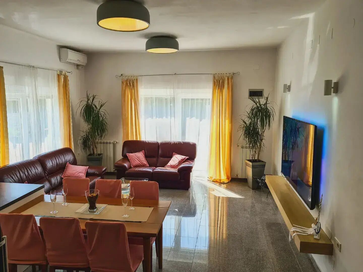 A modern living room with a brown leather sofa, a dining table with chairs, and a flat-screen TV mounted on the wall.