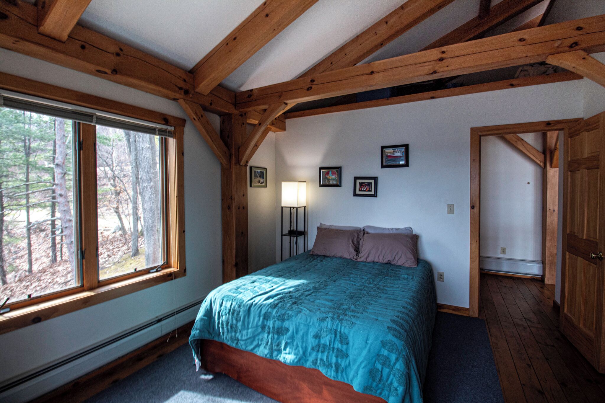 A bedroom with a large window, wooden beams, a bed with a blue bedspread, a standing lamp, and framed pictures on the wall.