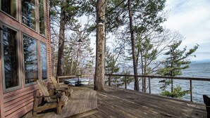 A wooden deck with Adirondack chairs, a railing, and a view of a lake and forest. - Architects Dream Home Overlooking Picturesque Lake Champlain (Keeseville)