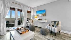 A spacious living room with a large sliding glass door leading to a balcony, a comfortable seating area with two armchairs and ottomans, a white console table with decorative items, and a beach-themed painting on the wall. - 🐟Spacious Balcony-Across from Beach-2 Pools-Cassine Way 303🐟 (Santa Rosa Beach)