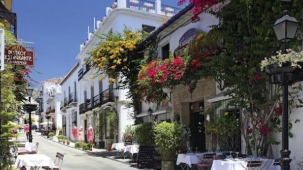 Una calle flanqueada por edificios blancos, algunos con balcones y flores colgantes. Hay mesas y sillas afuera, lo que sugiere terraza.