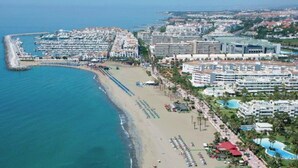 Vista aérea de una ciudad costera con un puerto deportivo, hoteles frente a la playa y un paseo marítimo.