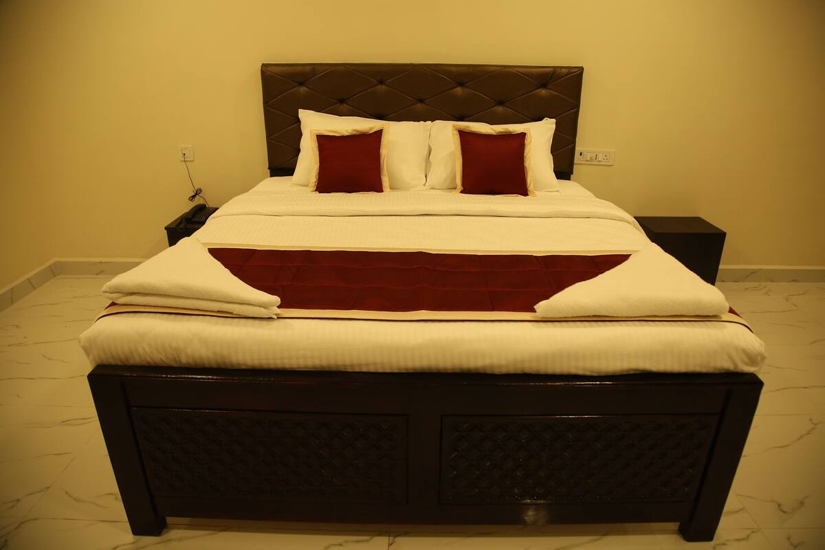 A neatly made bed with a dark headboard and two red pillows, a bedside table, and a power outlet on the wall.