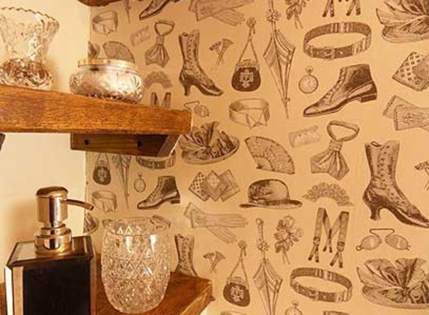 A room with a patterned wallpaper featuring various footwear designs, a wooden shelf with a crystal vase, and a metal object on the table.