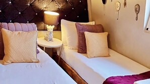 Hotellihuone, jossa on kaksi sänkyä, pyöreä pöytä lampun kanssa sekä koristeellisia seinäkoristeita.