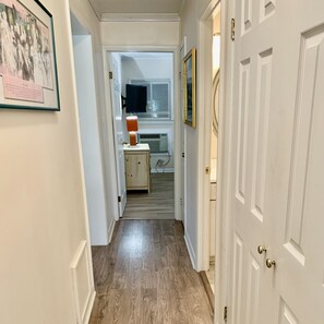 Un pasillo con piso de madera, una puerta a la derecha y una habitación con televisor y lámpara al fondo.