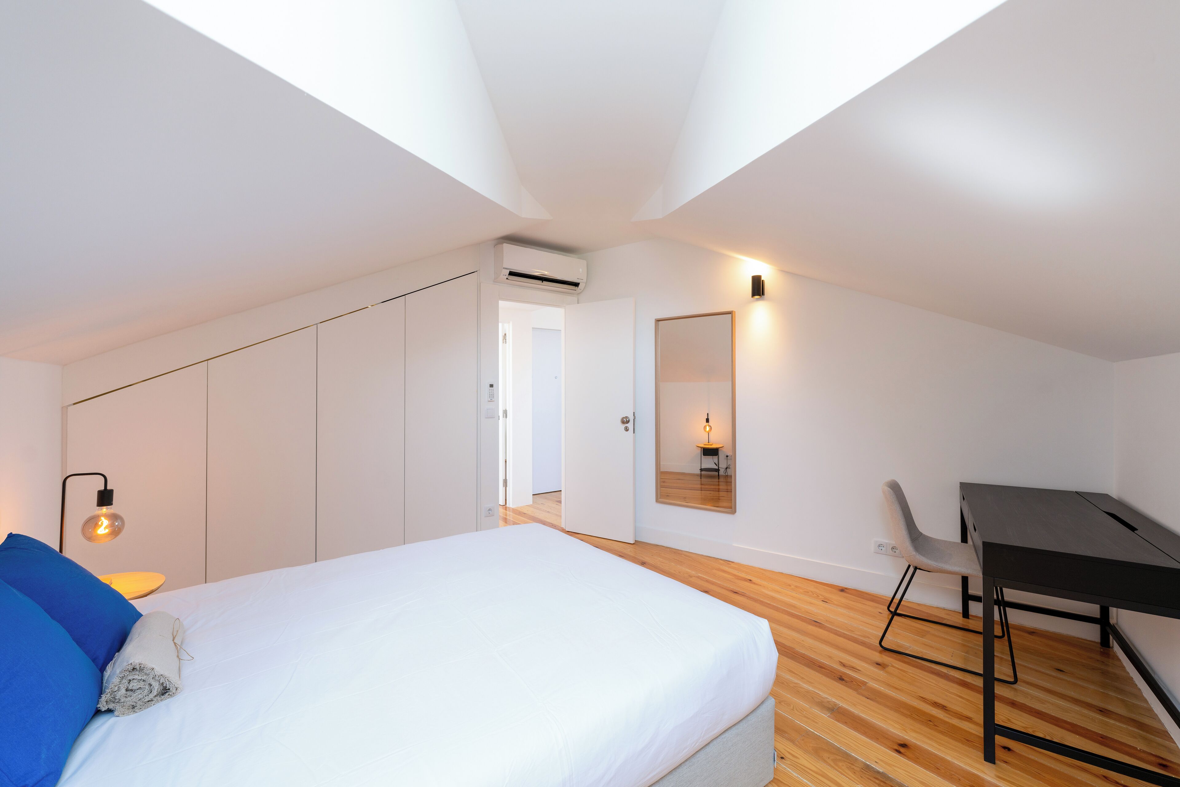 A modern bedroom with a bed, a desk, and a chair. There is a skylight above the bed and a wall-mounted mirror.
