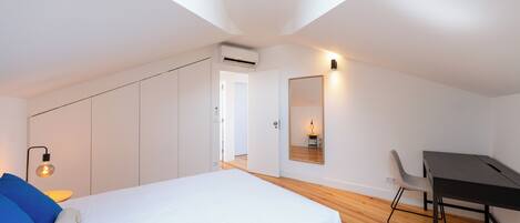 A modern bedroom with a bed, a desk, and a chair. There is a skylight above the bed and a wall-mounted mirror.