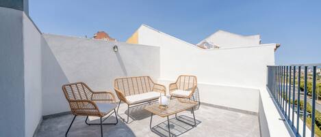 A balcony with two wicker chairs and a small table, a clear blue sky, and a white wall.