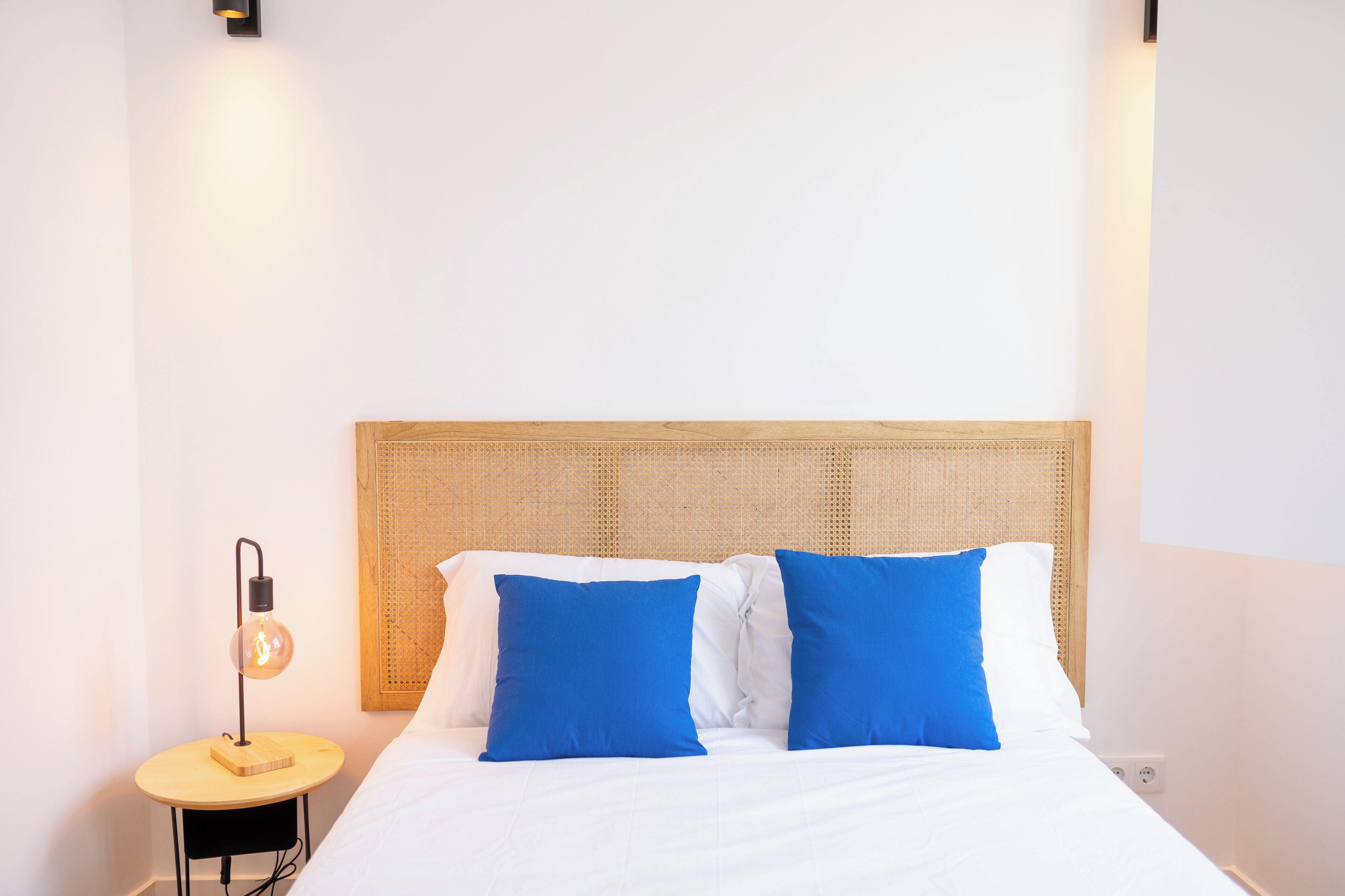 A neatly made bed with white and blue pillows, a round wooden side table with a lamp, and a woven headboard.