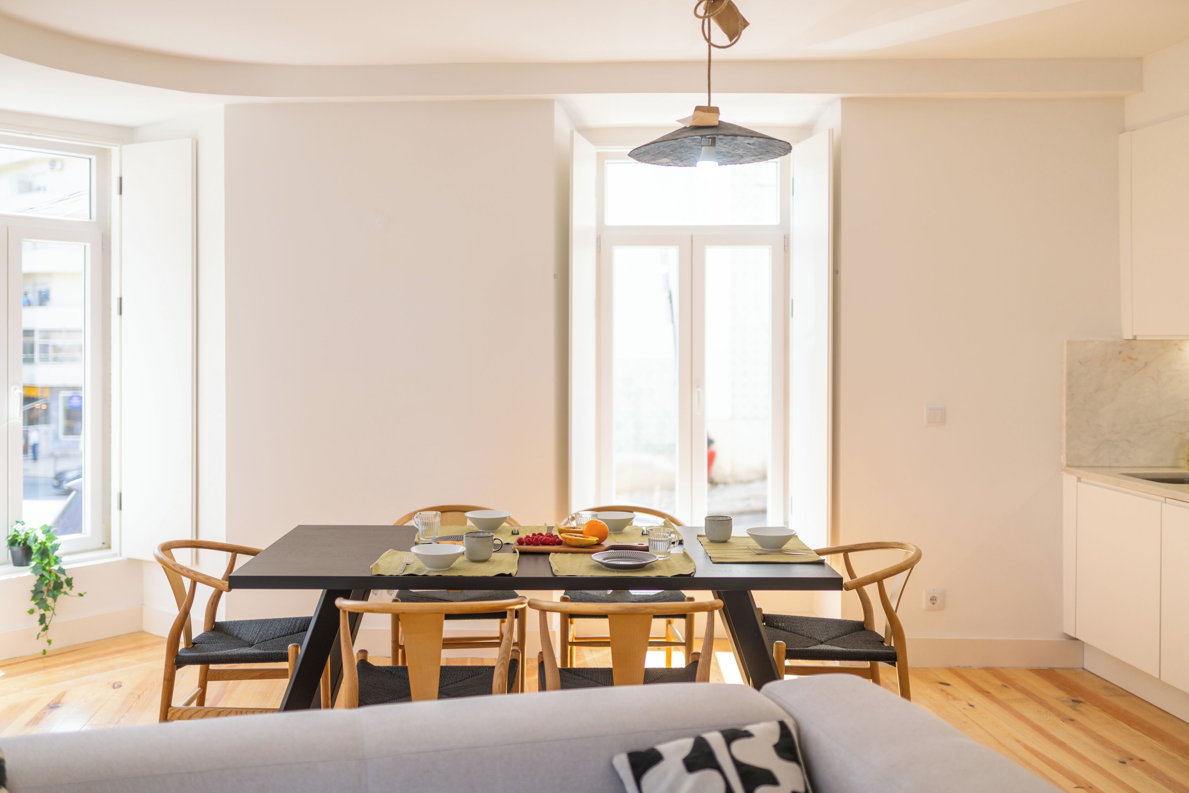 A dining area with a table set for four, surrounded by wooden chairs, a hanging light fixture, and a view through a large window.