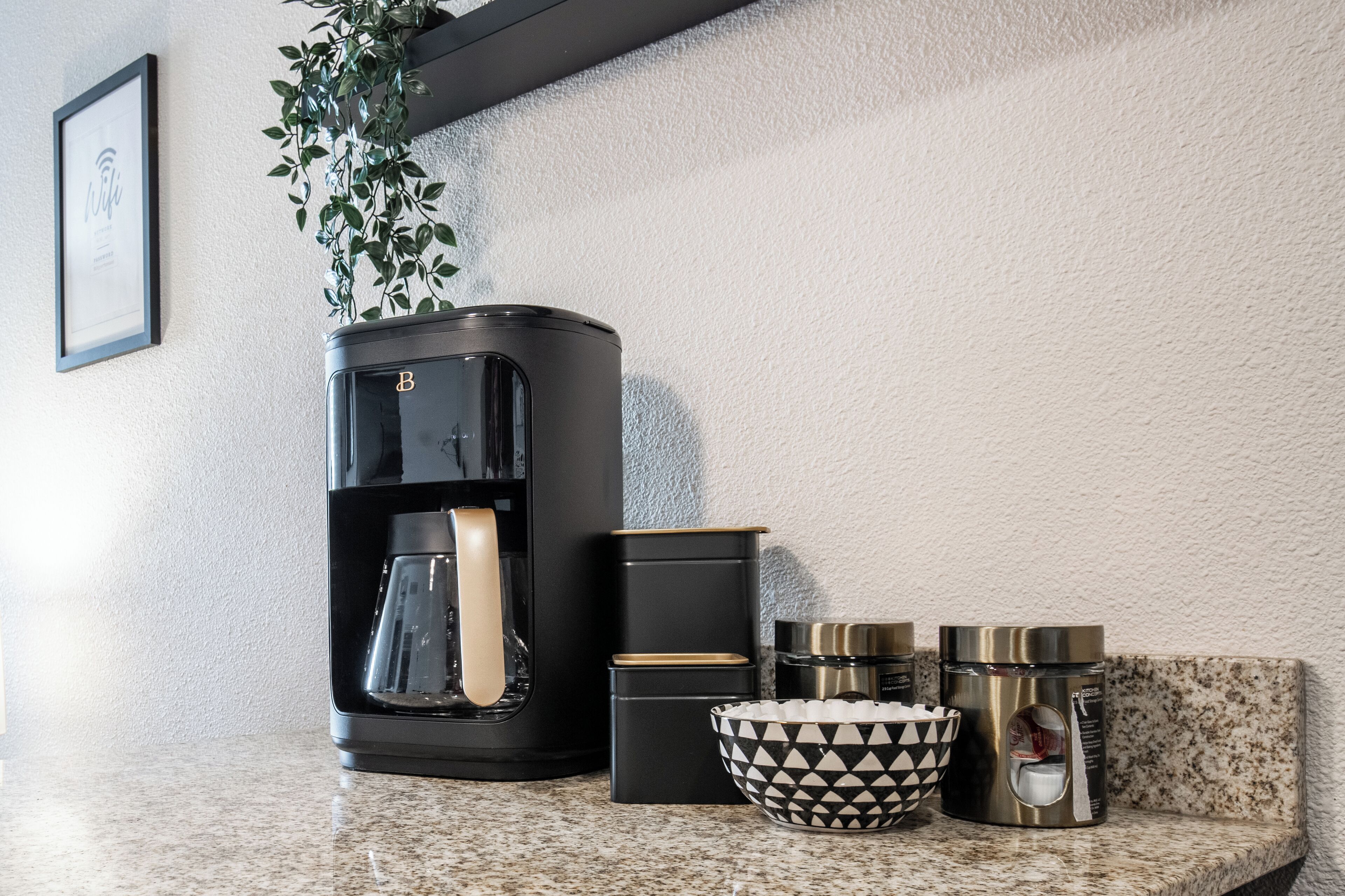 Una encimera de cocina con una cafetera, un tazón estampado en blanco y negro, y recipientes de acero inoxidable.