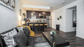 Een moderne woonkamer met een donker houten salontafel, een grijze bank met gekleurde kussens en een zwart tapijt. Op de achtergrond is een keuken zichtbaar met donker houten kastjes, een roestvrijstalen koelkast en een bar.
