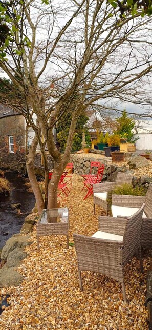 Relax in a serene outdoor setting surrounded by trees and a body of water, with comfortable wicker furniture and a glass table.
